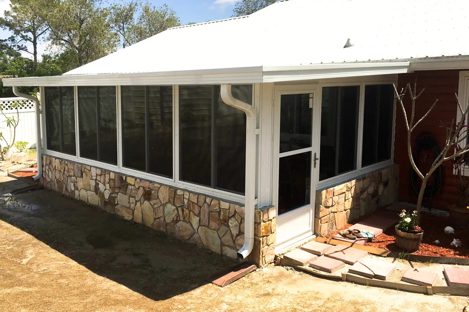 A white-framed glass sunroom built on a stone knee wall along the back of a single-storey home, with a screened entry door