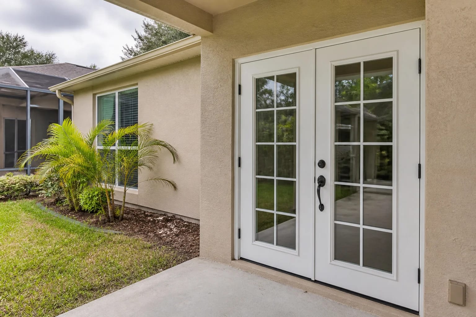 A white French door pair with colonial grids and dark bronze hardware set into the tan stucco wall of a Florida home, seen from a covered patio with areca palms and a screen enclosure alongside