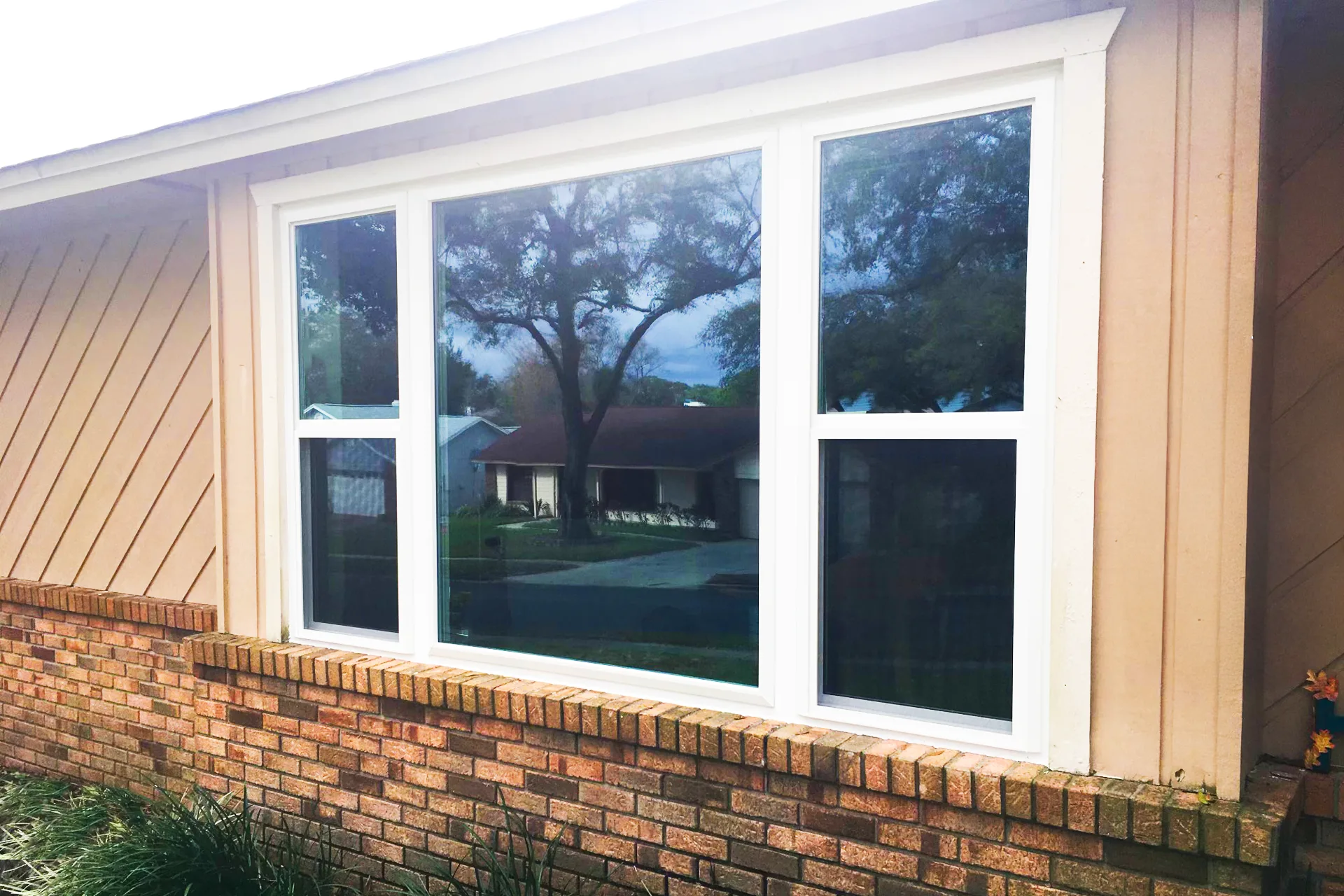Three new white vinyl replacement windows installed in a brick and board-and-batten Central Florida ranch home, reflecting a live oak in the front yard